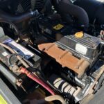 Close-up of a car engine bay showing a rusty exhaust manifold, battery, and assorted hoses and components