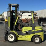 Green forklift in an outdoor industrial yard with pallets and equipment in the background under a clear blue sky, forks resting on the ground.