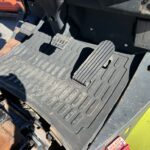 Driver footwell with textured rubber floor mat and three pedals (two small, one large) in an industrial vehicle; yellow body panel visible nearby.