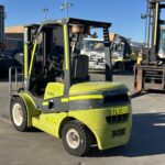Green forklift with yellow stripes parked in an outdoor industrial yard with other heavy equipment in the background.