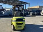 Front view of a lime-green forklift in an outdoor industrial yard with large metal bins behind it under a covered area.