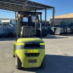 Front view of a lime-green forklift in an outdoor industrial yard with large metal bins behind it under a covered area.