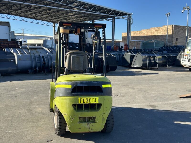 Front view of a lime-green forklift in an outdoor industrial yard with large metal bins behind it under a covered area.