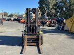 Front view of a rusty forklift parked in an outdoor industrial lot with orange equipment and fencing in the background.
