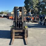 Front view of a rusty forklift parked in an outdoor industrial lot with orange equipment and fencing in the background.