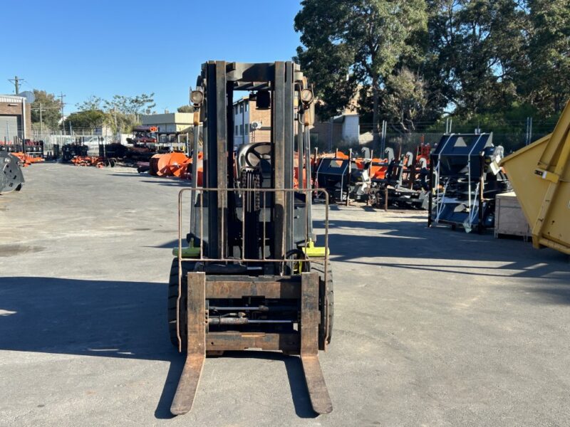 Front view of a rusty forklift parked in an outdoor industrial lot with orange equipment and fencing in the background.