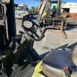 Close-up of a forklift cab interior with steering wheel, pedals, and control levers; industrial yard with forklifts and pallets in the background.
