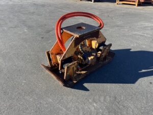 Old, rusted orange construction attachment with red hydraulic hoses coiled on top, sitting on a paved lot.