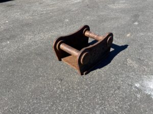 Rusty metal two-pin coupler attachment lying on asphalt, likely for heavy machinery.